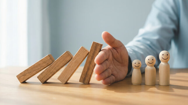 A businessperson uses a hand to stop falling wooden dominoes and protect smiling wooden peg figures conveying safety and teamwork