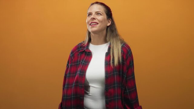Woman smiling and looking up with plaid shirt visible in studio, white tee and long hair on orange seamless backdrop, hands relaxed at sides; quiet joy.
