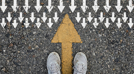 Person standing in worn white sneakers on textured asphalt facing a bold yellow forward arrow surrounded by many small white downward arrows evoking determination