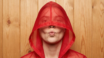 Playful woman wearing a sheer red hood over her face and puckering lips against warm wooden panel background creating a whimsical flirtatious mood