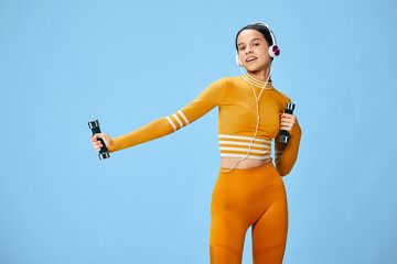 Young woman fitness in orange activewear holding small dumbbells and wearing headphones against a clean blue studio background, performing light strength exercises.