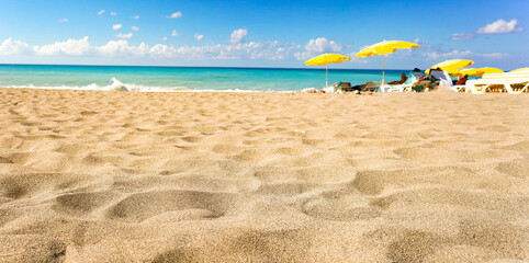 Low-angle perspective, detailed texture of sun-drenched sand. Blurry yellow umbrellas, turquoise...
