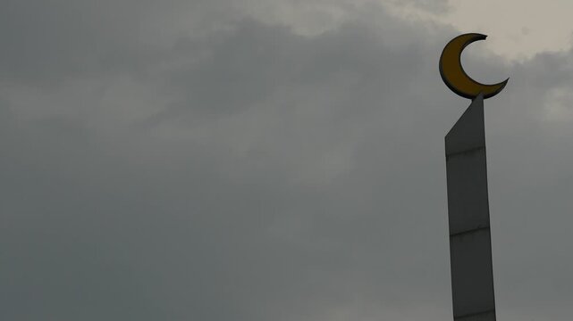 Time-lapse of a mosque minaret crescent symbol against a dramatic moving gray cloudy sky in landscape.