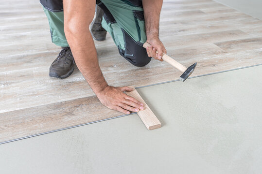 Floating floor work. The worker taps the board of vinyl plank to lock the click system. He is using hammer and wooden plank.