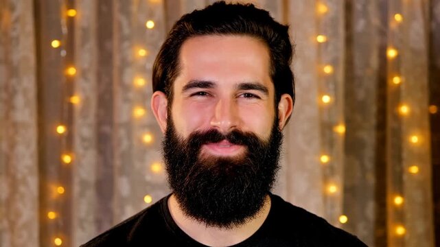 Handsome Middle Eastern man with full beard smiling and talking to camera against warm bokeh lights background in studio setting