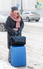 A pretty young woman in a hat and scarf stands with a blue suitcase and a black bag on a snowy road in the city in winter. Selective focus