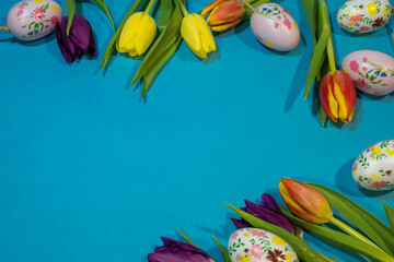 a festive border of yellow, orange, and purple tulips mixed with floral-painted Easter eggs against a solid blue background, creating a central copy space.