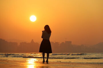 Silhouette of woman standing with smartphone on sea beach on city background. Sunrise on a coast, travel and holiday concept