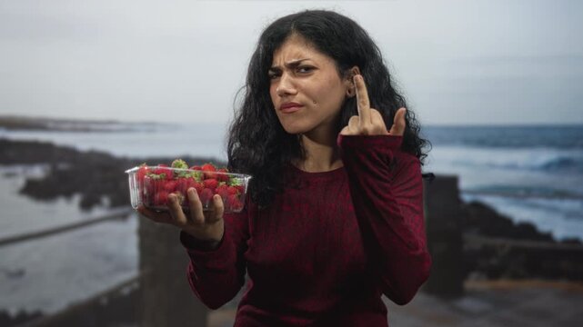 Young woman holds a clear plastic container of strawberries and raises middle finger in studio by the coast; defiance offering.