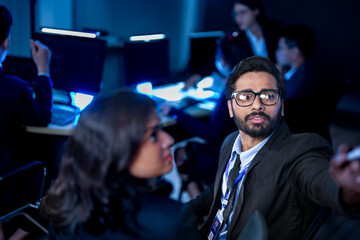 A passionate and visionary business leader with glasses presents a new strategy, pointing towards a large screen during a late-night meeting to inspire and guide his focused team.