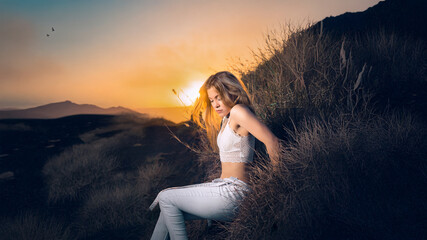 Latin American female model and singer reclining on volcanic slope at sunset, white jeans and crop top