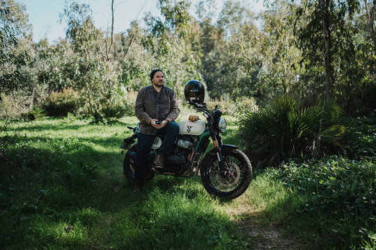 Hombre aventurero con barba en moto estilo scrambler en el bosque en dia soleado