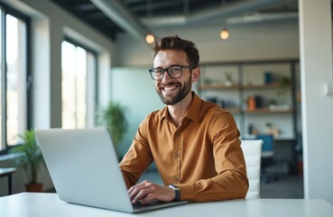 Fototapeta premium Man sits at desk working with laptop in office. He wears glasses, button down shirt and smiles. Professional uses notebook for work and studies at coworking workplace.