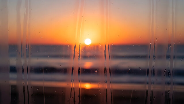Warm golden sunset over the tranquil ocean, seen through a steamy window with rain streaks and droplets. - Powered by Adobe