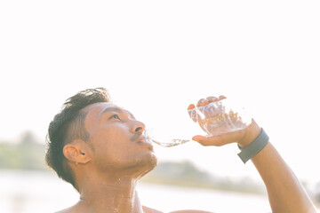 A young Asian man drinks water after exercising in a park during the summer, demonstrating an active and healthy lifestyle.