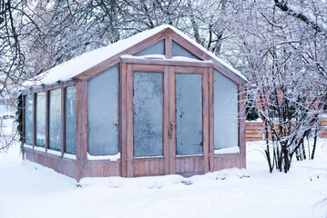 A wooden garden greenhouse covered in snow and frost, surrounded by icy winter trees in a backyard.