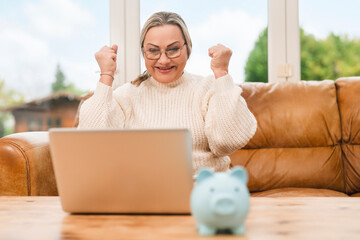 Woman celebrates on a couch with a laptop and a piggy bank, expressing joy during a financial moment