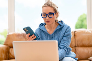 Young woman uses laptop and phone while sitting on couch in bright room during daytime