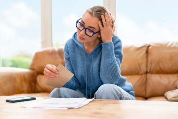 Woman sitting on couch looking at documents with concern during daytime in living room