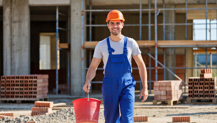Construction worker in blue overalls smiling on building site with red bucket