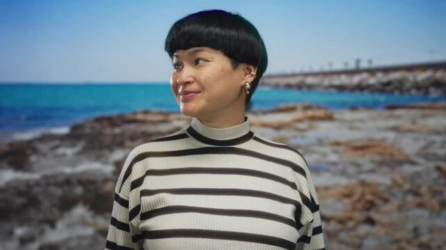 Smiling young woman standing on a rocky seaside with turquoise water backdrop, wearing striped shirt and showcasing joyful expression on a sunny beach day.