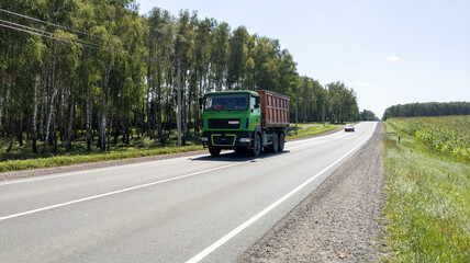 Dump truck drives along a road past a farm field and forest. Construction equipment drives through the countryside