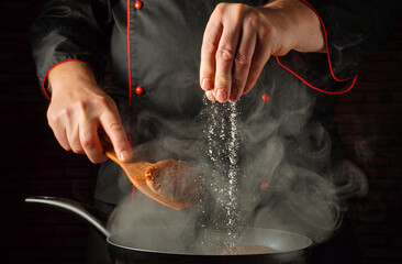 A chef stands in a kitchen, stirring food in a pan. He adds seasoning from a spoon into the pan, creating steam. The focus is on his hands and the blending of flavors