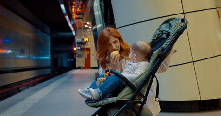 Young mother feeds her baby an apple while waiting for the subway train