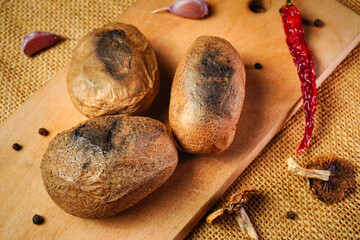 Three brown potatoes sit on a wooden cutting board. Nearby are garlic cloves, a red chili pepper, and dried mushrooms. The scene captures a kitchen setting with natural textures