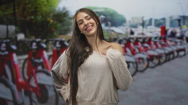 Teenage girl touches shoulder and poses beside a row of rental bicycles on street; confidence youth.