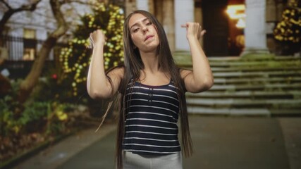 Young woman with long hair and braces reaches forward with both hands and outstretched palms on stone steps of building near lit trees at night; defiance rebellion.