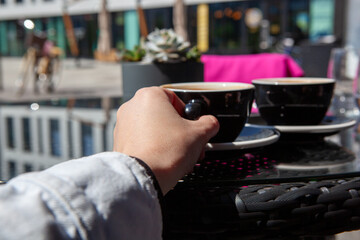 A person holds a coffee cup while sitting at a table outside. There are two cups on the table. The setting shows buildings and outdoor decor under sunlight