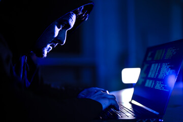 Mysterious hacker in black hoodie stealing confidential data on laptop. Cyber criminal coding dangerous malware virus to attack network system in dark blue room.