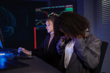 Professional female analysts discussing global market trends. Businesswoman pointing at financial data on computer screen while colleague works in night office.