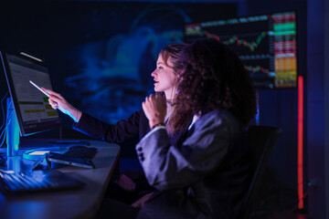 Dedicated business team working late on corporate project. Professional women analyzing digital performance data on screens in modern dark blue office at night.
