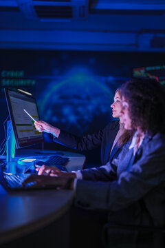 Cyber security specialists monitoring global network threats. Female tech expert pointing at digital system data on screen while partner analyzing code at night.