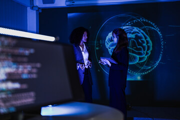 Two female researchers discussing neurology near digital brain projection. Scientists analyzing artificial intelligence data and mental health concepts in dark blue lab.