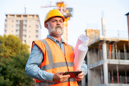 A construction worker wearing an orange vest and hard hat stands confidently at an urban site, holding blueprints