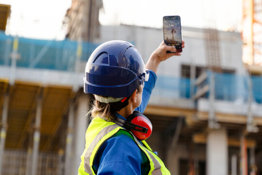 Female construction worker in safety gear observes ongoing work at construction site, focusing on crane lifting materials under clear skies