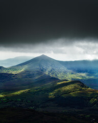 This is a stunning view of the rugged mountain peaks of Ireland. Dark storm clouds loom overhead, while patches of sunlight illuminate the lush green valleys below.