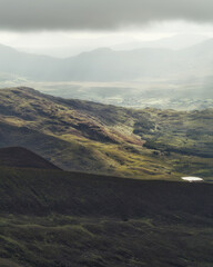 Mountains and a valley are visible in County Kerry, Ireland. The sky is overcast, and there is a small lake in the distance, showcasing the landscape's beauty.