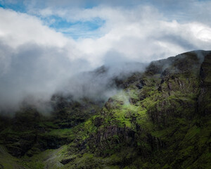 Mist envelops a verdant mountain, partially concealing its rocky face under a blanket of ethereal clouds and a peek of blue sky above.