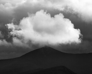 Cumulus cloud hovers just above the peak of a silhouette mountain on a stormy day. The sky is dark and full of clouds. The landscape appears dark and desolate.