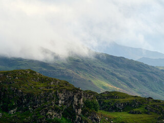 Misty mountains rise above rugged cliffs and green vegetation under soft natural light.