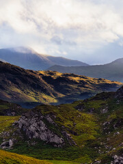 Rolling green hills are bathed in soft light, creating shadows and highlights across the Irish landscape. Clouds hang over distant peaks in Ireland.