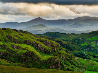 A green hillside covered in vegetation and rocky outcroppings slopes down into a valley. Distant mountains rise in the background under a cloudy sky in National Park.