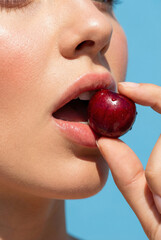 Close-up of a woman's sensual lips holding a fresh red cherry. Perfect skin on a clean blue background. Concept of beauty, temptation, and health.