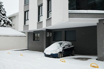 Snow covered car parked beside a modern residential building in winter. Urban lifestyle scene showing cold weather, daily routine, and residential architecture.