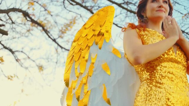 Greek goddess fantasy happy woman folds hands in prayer. White golden wings costume angel dress metal gold jewels crown tiara. Sexy girl angel style. Summer nature divine magic sun light sky trees