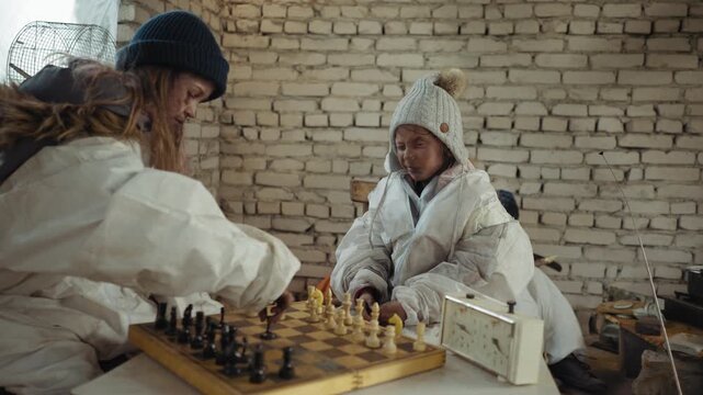 Two orphans in dirty winter clothes play chess on wooden board as older girl taps clock after pawn move while someone partially visible in background sharpens wood inside cold brick walled shelter